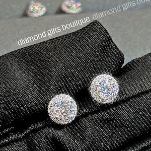 May include: A pair of halo round earrings with a central stone of 0.5 ct and 44 side stones. The earrings are held on a black fabric. The earrings measure 7.4 x 7.4mm. The text "diamond gifts boutique" is visible.