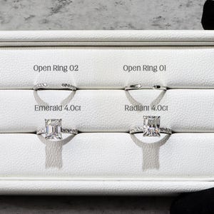 May include: A white display case holding four diamond rings. The top two rings are open-style designs. Below are two rings, one with an emerald cut diamond and the other with a radiant cut diamond, both labeled 4.0ct. The case is held by a black-gloved hand.