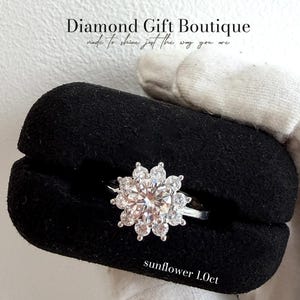 May include: A silver ring featuring a sunflower-shaped diamond, with a 1.0ct centre stone. The ring is presented on a black velvet box. The text "Diamond Gift Boutique" and "sunflower 1.0ct" are visible.