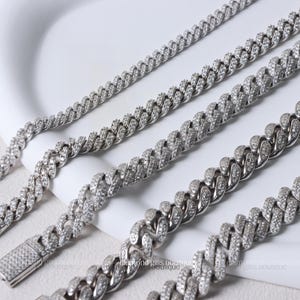 May include: A selection of silver-coloured, diamond-set chain necklaces displayed on a white surface. The necklaces are arranged diagonally. The chains feature a curb link design, with each link covered in small, sparkling diamonds. Clasps are visible on some of the necklaces.