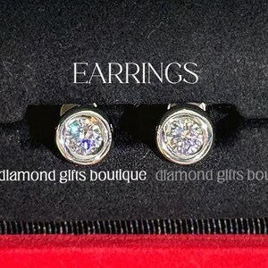 May include: A diamond pendant necklace and a pair of diamond stud earrings are presented in a red jewellery box. The necklace features a single round diamond in a bezel setting. The earrings also have round diamonds. The image includes the words "NECKLACE" and "EARRINGS".