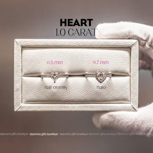 a hand holding a white jewelry box containing two diamond rings. The text above the box reads "HEART 1.0 CARAT".