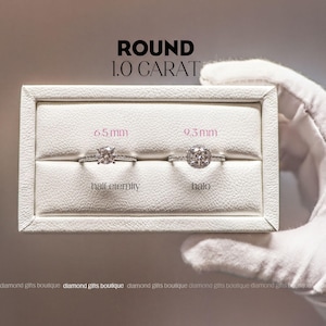a hand holding a white jewelry box containing two diamond rings, one labeled "half eternity" and the other labeled "halo". The box is placed on a white surface, and the background is blurred.