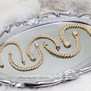May include: Several gold-colored, diamond-studded bracelets are displayed on a silver mirrored tray. The bracelets are arranged in a curved pattern, showcasing their intricate design and clasp details. The tray sits on a fluffy white surface.