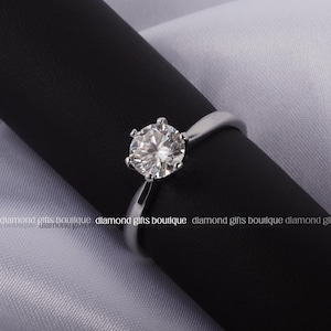 May include: A silver engagement ring with a large, round, clear diamond set on a simple band. The ring is displayed on a black surface against a white background. The diamond sparkles, catching the light.