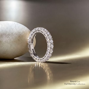 May include: A silver-toned full eternity ring featuring a continuous band of round-cut clear stones set in a prong setting. The inner band is clearly engraved with the text S925. The text Round Cut Full Eternity Collection is visible.