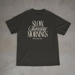 May include: Dark gray t-shirt with the text "SLOW Sunday MORNINGS" in a stylized font. Below the text is "GOOD VIBES ONLY" in a smaller font. The shirt is made of a soft material.