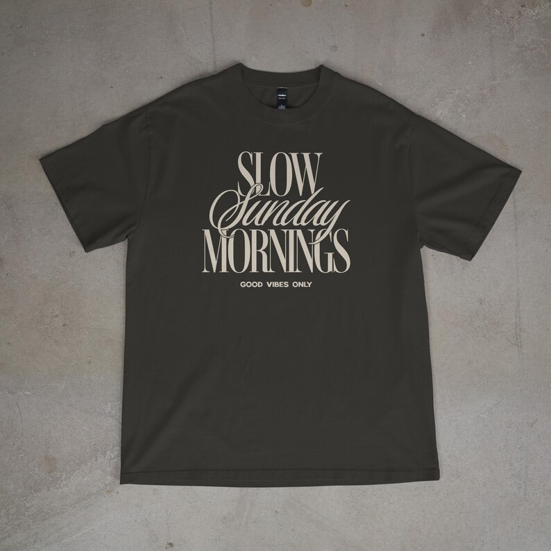 May include: Dark gray t-shirt with the text "SLOW Sunday MORNINGS" in a stylized font. Below the text is "GOOD VIBES ONLY" in a smaller font. The shirt is made of a soft material.