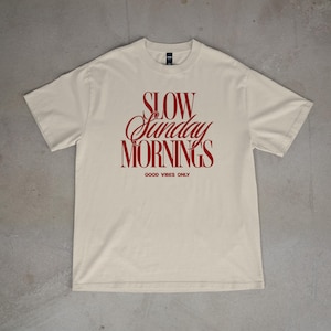 May include: A cream-colored t-shirt with the words "SLOW Sunday MORNINGS" in a red, cursive font. Below the text is "GOOD VIBES ONLY" in a smaller, block font. The shirt is laid flat on a gray surface.