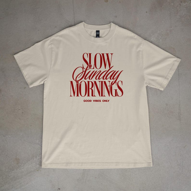 May include: A cream-colored t-shirt with the words "SLOW Sunday MORNINGS" in a red, cursive font. Below the text is "GOOD VIBES ONLY" in a smaller, block font. The shirt is laid flat on a gray surface.
