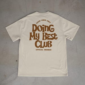 May include: Cream-colored t-shirt with the text "GOOD VIBES ONLY" above "Doing My Best Club" in brown, retro-style lettering. The words "OFFICIAL MEMBER" are printed below the club name. The shirt is displayed on a gray surface.