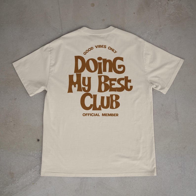 May include: Cream-colored t-shirt with the text "GOOD VIBES ONLY" above "Doing My Best Club" in brown, retro-style lettering. The words "OFFICIAL MEMBER" are printed below the club name. The shirt is displayed on a gray surface.