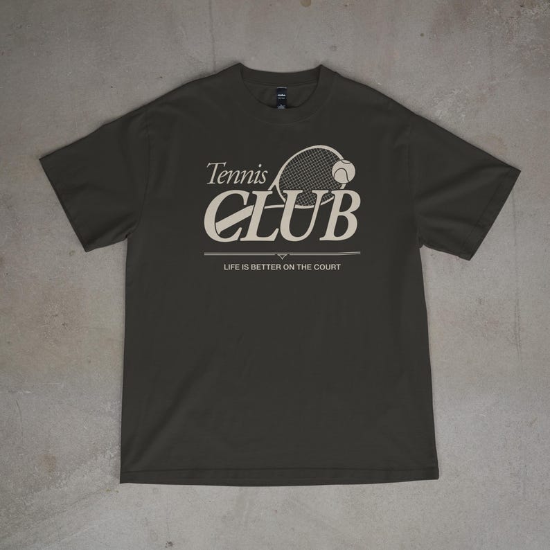 Op de afbeelding: Donkergrijs t-shirt met de tekst "Tennis CLUB" in cr&egrave;mekleurige letters, met een tennisracket en bal afbeelding. De tekst "LIFE IS BETTER ON THE COURT" staat eronder.