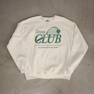 Op de afbeelding: Cr&egrave;mekleurige sweatshirt met een groene tennisracket en bal afbeelding. De tekst luidt "Tennis Club" boven "CLUB" en "LIFE IS BETTER ON THE COURT" eronder. Een casual, sportief ontwerp.