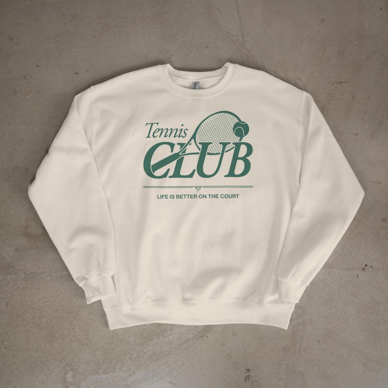 Op de afbeelding: Cr&egrave;mekleurige sweatshirt met een groene tennisracket en bal afbeelding. De tekst luidt "Tennis Club" boven "CLUB" en "LIFE IS BETTER ON THE COURT" eronder. Een casual, sportief ontwerp.