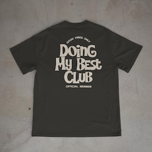 May include: Dark gray t-shirt with the text "GOOD VIBES ONLY" and "DOING MY BEST CLUB OFFICIAL MEMBER" in a retro font. The shirt is laid flat, showcasing the design on the back.