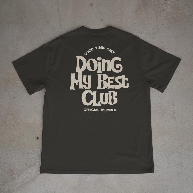May include: Dark gray t-shirt with the text "GOOD VIBES ONLY" and "DOING MY BEST CLUB OFFICIAL MEMBER" in a retro font. The shirt is laid flat, showcasing the design on the back.
