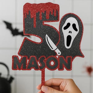 May include: A glittery red and black cake topper with the number 5, a knife, and a ghost face. The name "MASON" is below the number. The topper is held by a hand. A bat silhouette is in the background.
