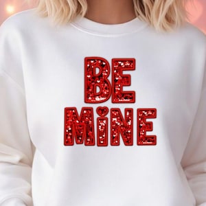 May include: A white sweatshirt with the text "BE MINE" in red, heart-filled letters. The sweatshirt is worn with blue jeans. The background includes a floral arrangement and heart-shaped decorations, suggesting a romantic theme.
