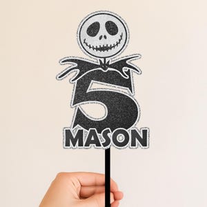 May include: A black and silver glitter cake topper featuring Jack Skellington's head, the number 5, and the name MASON. The topper is held by a black stick, perfect for a birthday celebration.