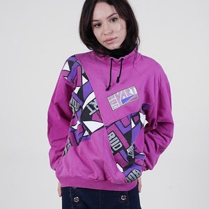 Könnte beinhalten: Eine Frau trägt einen rosa Pullover mit geometrischem Muster und blaue Jeans. Der Pullover hat einen hohen Kragen und einen Kordelzug.