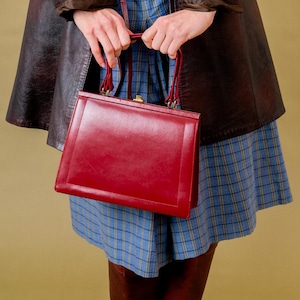 May include: A vintage-style outfit featuring a brown leather coat, a red handbag, a blue plaid skirt, and burgundy knee-high boots. The coat has a classic design, and the handbag has a structured shape. The outfit is completed with a cream-colored top.