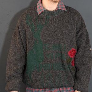 May include: A gray sweater with a green deer and red roses embroidered on the front. The sweater has a crew neck and long sleeves. The person is wearing a plaid shirt underneath the sweater and black pants.