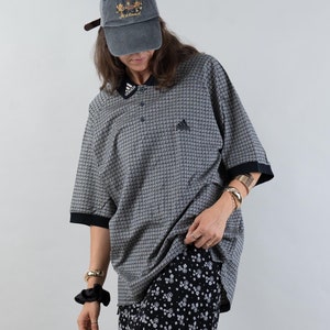 Op de afbeelding: Een persoon draagt een grijs en zwart geruit poloshirt met een zwarte bloemenrok en witte sneakers. De persoon draagt een grijze baseballpet met de tekst "Royal" erop geborduurd.