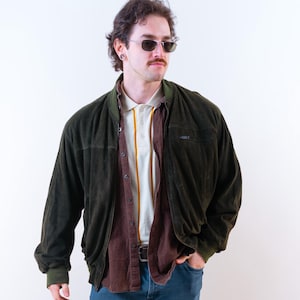 Puede incluir: Una persona lleva una chaqueta bomber de pana verde oscuro sobre una camisa abotonada marrón y una camiseta polo blanca con rayas amarillas. La persona también lleva vaqueros azules y zapatos negros.