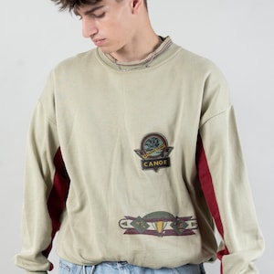Könnte beinhalten: Ein hellgrüner Langarm-Pullover mit roten Akzenten an den Ärmeln. Der Pullover hat einen grafischen Aufdruck eines Globus mit dem Wort "CANOE" darunter. Das Model trägt eine blaue Jeans-Shorts.