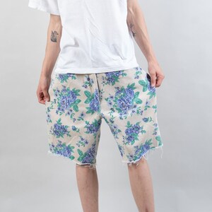 May include: A person wearing a white t-shirt and beige floral print shorts. The shorts have a frayed hem.