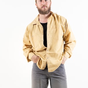 Könnte beinhalten: Eine Person trägt ein gelbes Hemd mit Knöpfen, eine graue Hose und eine braune Baseballkappe. Sie steht mit den Händen in den Taschen.