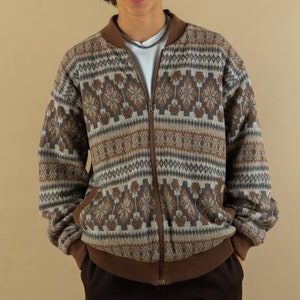 May include: A man wearing a brown and white patterned cardigan with a zipper closure. He is also wearing a green baseball cap with a white logo that says "Mountain", a white t-shirt, and brown pants.