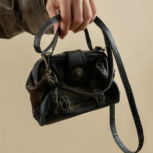 Könnte beinhalten: Eine schwarze Ledertasche mit braunem und schwarzem Muster. Die Tasche hat einen langen Riemen und einen Klappverschluss mit einer Metallschnalle.