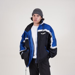 May include: A blue and black color block jacket with white accents and a zipper closure. The jacket has a hood and is worn over a white t-shirt. The person is wearing black pants and black boots.
