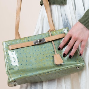 May include: A light green handbag with a textured finish and tan leather accents. The rectangular bag has a silver clasp and a matching tan leather tag. The bag is held by tan leather handles.