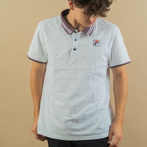Op de afbeelding: Een wit poloshirt met een bordeauxrode en marineblauwe bies rond de kraag en mouwen. Het shirt heeft een klein geborduurd logo op de borst met "FILA" in rood en blauw.