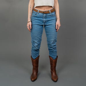 Peut inclure: Une femme porte un haut court blanc &agrave; manches courtes, un jean bleu, une ceinture marron et des bottes de cowboy marron. Elle porte &eacute;galement des lunettes de soleil. Le fond est gris uni.