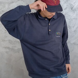 May include: A navy blue long-sleeved polo shirt with a small embroidered design on the chest. The shirt has a button-up collar and a button placket. The person is wearing blue jeans and a red and black baseball cap with the text "Chicago Bulls" on it.