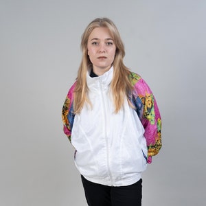 Puede incluir: Una mujer con una chaqueta blanca con estampado de mariposas rosa, amarillo y azul en las mangas. Lleva pantalones negros y zapatillas verdes.