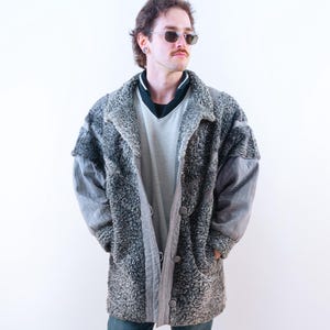 Könnte beinhalten: Ein grauer, strukturierter Mantel mit Knopfleiste und großem Kragen. Der Mantel wird mit einem hellgrauen Hemd, dunkelblauen Jeans und schwarzen Schuhen getragen. Der Mantel hat eine einzigartige, gemusterte Textur.