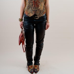 Könnte beinhalten: Eine Person trägt eine schwarz-goldene Blumenweste, eine schwarze Lederhose und gemusterte Schuhe. Sie trägt auch eine Sonnenbrille und hält eine rote Handtasche. Das Outfit hat eine Vintage- und Bohème-Ästhetik.