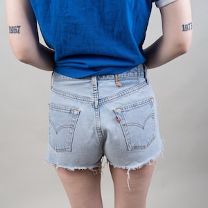 Può includere: Una persona indossa una maglietta blu con bordini bianchi e pantaloncini di jeans blu chiaro. La persona ha dei tatuaggi sulle braccia che dicono "1967" e "1973". Indossa scarpe da ginnastica bianche con accenti neri.