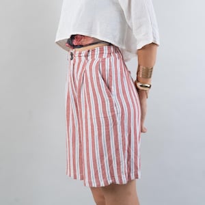 Könnte beinhalten: Eine Frau trägt ein weißes, bauchfreies Oberteil mit einem weißen Schal, rot-weiß gestreifte Shorts und goldene Armbänder. Sie trägt eine Sonnenbrille.