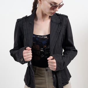 May include: A black pinstriped blazer featuring epaulets on the shoulders, a notched lapel, and a buckled belt at the waist. The jacket is worn over a black top with a floral pattern and paired with light tan trousers, complemented by aviator sunglasses.