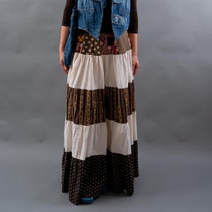 Könnte beinhalten: Eine Frau trägt ein braunes Langarmshirt, eine Jeansweste und einen langen Patchwork-Rock. Der Rock hat abwechselnd weiße, braune Blumen- und braune Polka-Dot-Stoffbahnen. Eine farbenfrohe Perlenkette und Sonnenbrille vervollständigen den Look.