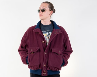 May include: A burgundy bomber jacket with a blue ribbed collar and cuffs. The jacket has a zipper closure and two front pockets. The model is wearing black pants and brown shoes.