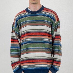 Könnte beinhalten: Ein mehrfarbiger gestreifter Pullover mit Rundhalsausschnitt. Der Pullover hat horizontale Streifen in Rot-, Orange-, Grün-, Blau- und Weißtönen. Die Manschetten und der Ausschnitt sind einfarbig marineblau. Die Person trägt eine dunkelblaue Cordhose.