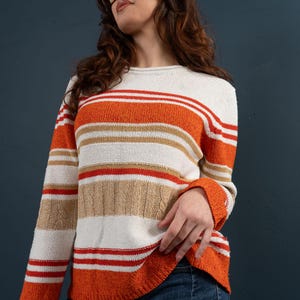 Puede incluir: Un suéter de manga larga con rayas horizontales en naranja, blanco, rojo y beige. El suéter tiene un cuello redondo y un diseño de punto trenzado en la parte inferior. Los puños y el dobladillo son naranjas.