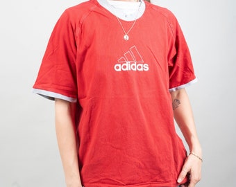 May include: A red t-shirt with a white Adidas logo and a white trim around the neck and sleeves. The model is wearing a grey baseball cap with a white NY logo.